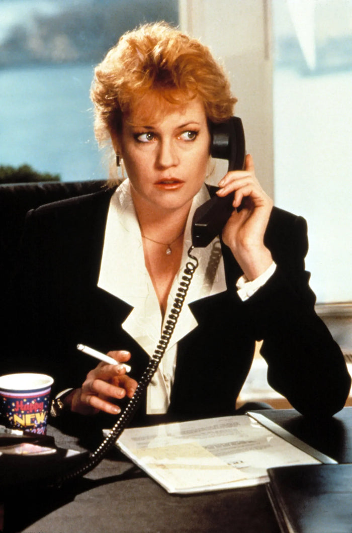 Melanie Griffith in the 1988 film Working Girl, wearing a black power suit with white lapels, holding a phone and cigarette—an iconic image of 80s office fashion and power dressing.